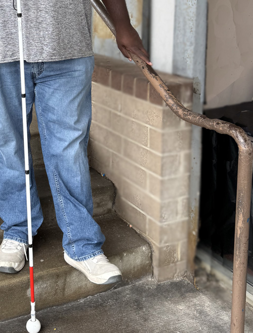 Photo shows a man wearing blue jeans with a white cane standing on the bottom step of a stairway, with his hand on the railing.  The railing extends another 8 inches beyond his hand.